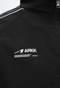 Μαύρο μπουφάν με λευκό φερμουάρ, λευκό περίγραμμα στον ώμο και λευκό κείμενο "ARKK Copenhagen Essentials Innovation Included Est 2015" στο στήθος.