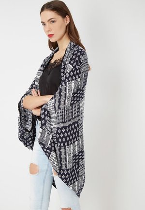 Mujer con cabello castaño que viste una blusa de encaje negra, jeans rotos blancos y un chal azul marino con estampado, de pie con los brazos cruzados sobre un fondo blanco.