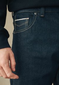 Los pantalones vaqueros de mezclilla oscura presentan un ajuste slim, costuras visibles, un bolsillo frontal con un acento más claro y un detalle de botón metálico.