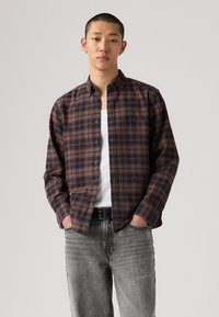 Καφέ και μπλε καρό πουκάμισο με κουμπιά από ελαφρύ ύφασμα, μακριά μανίκια, κλασικό γιακά, φορεμένο πάνω από λευκή αμάνικη μπλούζα, σε συνδυασμό με γκρι τζιν.