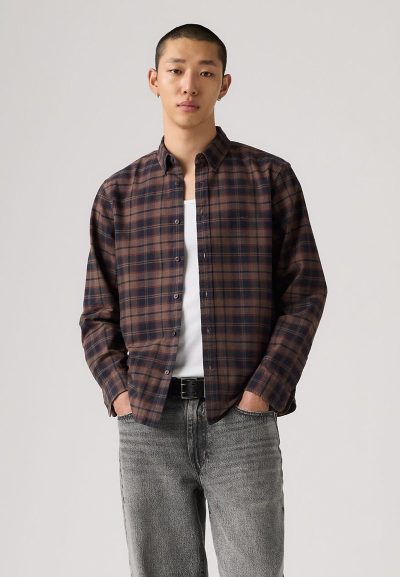 Καφέ και μπλε καρό πουκάμισο με κουμπιά από ελαφρύ ύφασμα, μακριά μανίκια, κλασικό γιακά, φορεμένο πάνω από λευκή αμάνικη μπλούζα, σε συνδυασμό με γκρι τζιν.