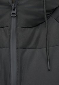 Fekete pufferszövet kabát magas gallérral, elöl cipzárral és állítható zsinórral. Simító, matt felületű, varrott panelokkal.