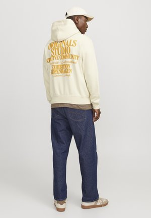 Cream hoodie with large orange lettering on the back, paired with dark blue jeans and beige sneakers, showing a relaxed fit silhouette.