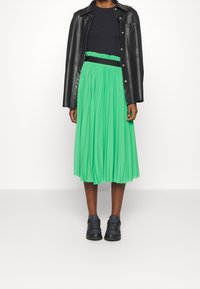 Jupe midi plissée verte avec une taille élastique, associée à un haut noir ajusté et une veste en cuir noire, complétée par des baskets noires.