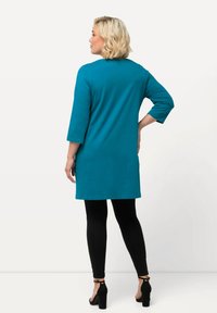 Teal tunika med trekvartsärmar, bakvy; kombinerad med svarta leggings och svarta klacksandaler. Slät, stretchig tyg.
