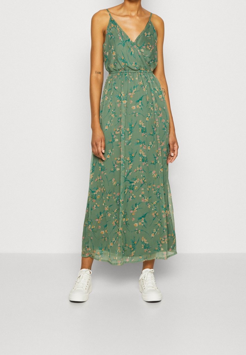 Robe maxi verte à fleurs sans manches avec fines bretelles, décolleté en V et taille froncée, portée avec des baskets blanches.