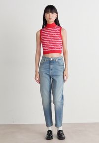 Top court rouge avec un texte blanc horizontal, associé à un jean taille haute bleu clair et des chaussures noires. Le modèle se tient devant un fond neutre.