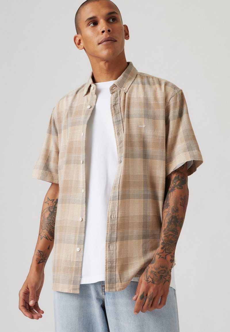 Short-sleeved, beige plaid shirt with button-down collar, checkered pattern, and light blue accents, worn over a white T-shirt.