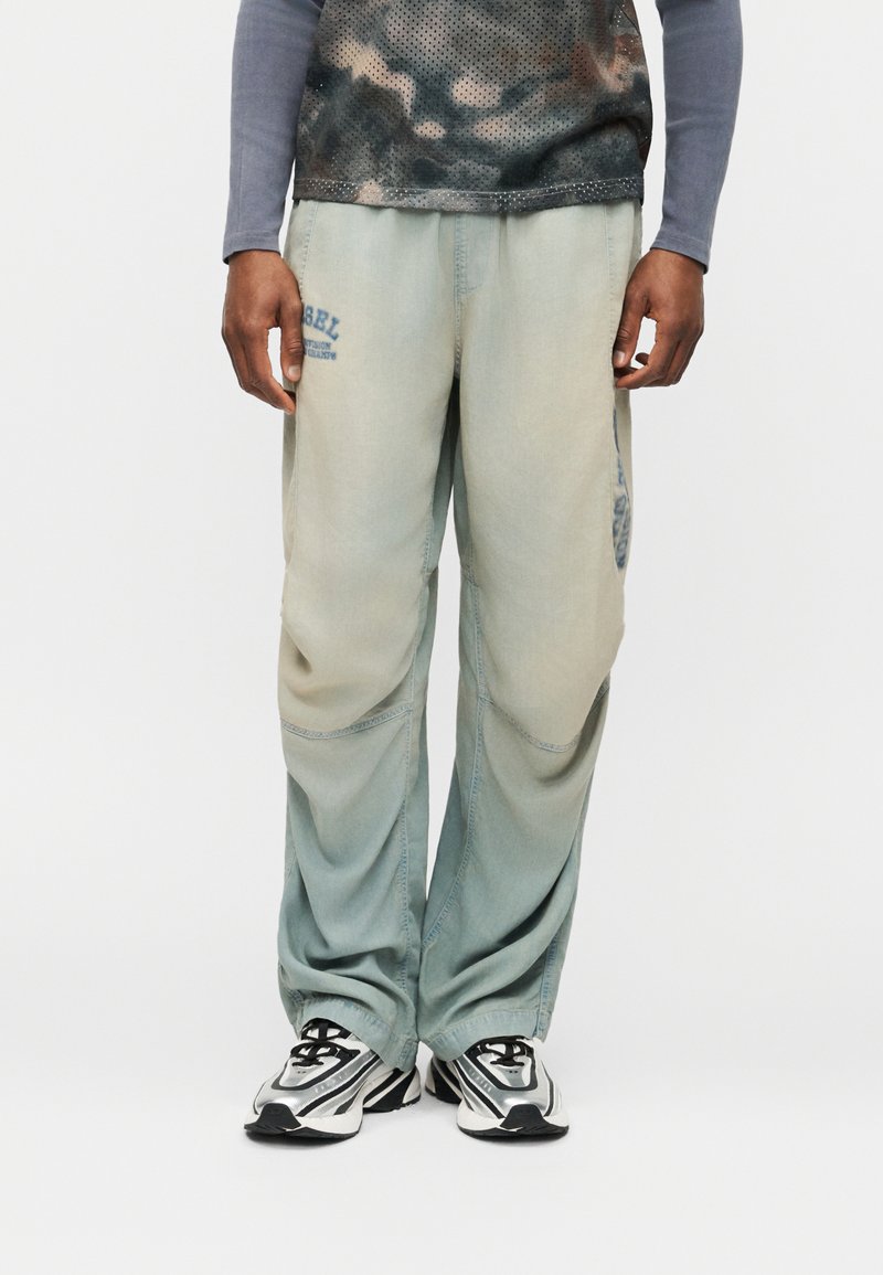 Άνδρας που φοράει φθαρμένο ανοιχτοπράσινο φαρδύ παντελόνι με κείμενο στο αριστερό μηρό και ασημί-λευκά αθλητικά παπούτσια, στέκεται μπροστά σε μια απλή φόντο.