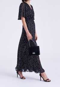 Svart maxi-klänning med vita polka prickar, korta fjäderärmar, djup V-ringning och cinchad midja, tillsammans med en svart texturerad handväska. Klacksandaler.