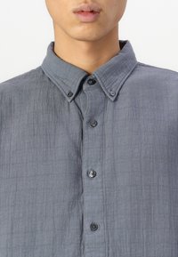 Camisa gris de botones, hecha de una tela texturizada con un aspecto arrugado. Presenta un collar puntiagudo y botones negros en el frente.