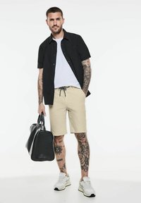 Męski strój składający się z czarnej koszuli z krótkim rękawem, białego t-shirtu, beżowych szortów i białych sneakersów. Trzyma czarną torbę podróżną.