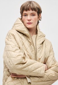 Μπεζ καπιτονέ μπουφάν με μεγάλο καπό, διαθέτει φερμουάρ μπροστά και υπερμεγέθη σιλουέτα. Το ύφασμα φαίνεται λείο και ελαφρώς γυαλιστερό.