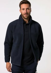 Chaqueta de forro polar navy con un cuello alto, cierre de cremallera en el frente y bolsillos laterales. Llevada sobre una camiseta polo oscura; presenta un logo sutil en el pecho.