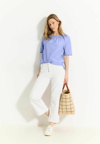 Blaues Leinenblusengewand mit gerafftem Ausschnitt, weiße weit geschnittene Hose und eine Tasche mit kariertem Muster. Weiße Sneaker runden das Outfit ab.