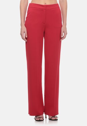 Donna che indossa pantaloni rossi a vita alta e gamba larga e tacchi neri aperti in piedi su sfondo bianco.