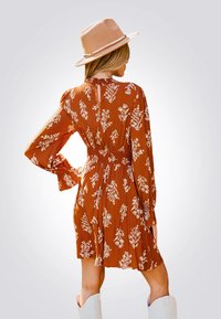 Vestido floral marrón con mangas largas, cintura ceñida y detalle de abertura en la espalda. Combinado con un sombrero de ala ancha de color claro y botas blancas.