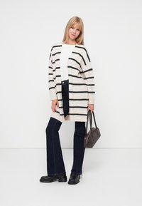 Gestreifter Strickcardigan in Creme und Schwarz, offene Front, lange Ärmel, kombiniert mit dunklen Jeans und schwarzen Knöchelbooties, dazu eine graue Handtasche.