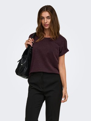 Femme aux cheveux bruns portant un haut court à manches courtes violet foncé et un pantalon noir, tenant un sac à main en cuir noir sur l'épaule.
