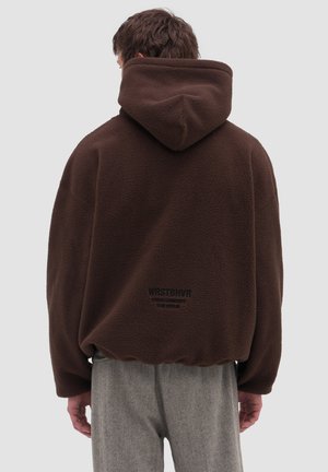 Mand iført en mørkebrun fleece-hoodie med hætten nede, grå bukser, stående med ryggen til på en ensfarvet lys baggrund.