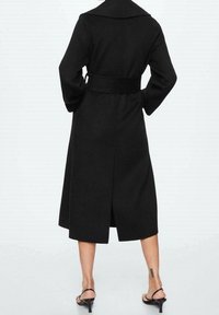 Manteau en laine noire avec un large col, des manches longues et une taille ceinturée. Le design comprend une fente au dos et une longueur mi-mollet.