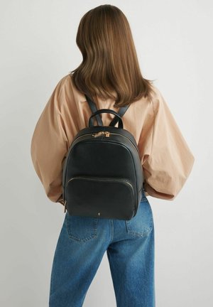 Osoba o brązowych włosach do ramion, ubrana w beżową bluzkę i niebieskie dżinsy, niosąca czarny skórzany plecak ze złotymi zamkami.