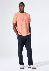 Koraalrood T-shirt met korte mouwen, een ronde hals en een subtiel logo, gecombineerd met marineblauwe broek en witte sneakers met beige accenten.