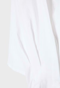 Tissu blanc avec des coutures visibles et des plis doux, probablement une partie d'un vêtement, sur un fond gris clair.