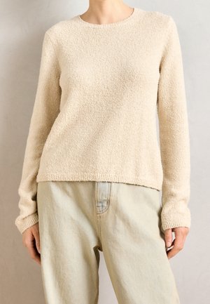 Personne portant un pull beige texturé à manches longues et un pantalon beige clair taille haute, debout devant un fond uni.