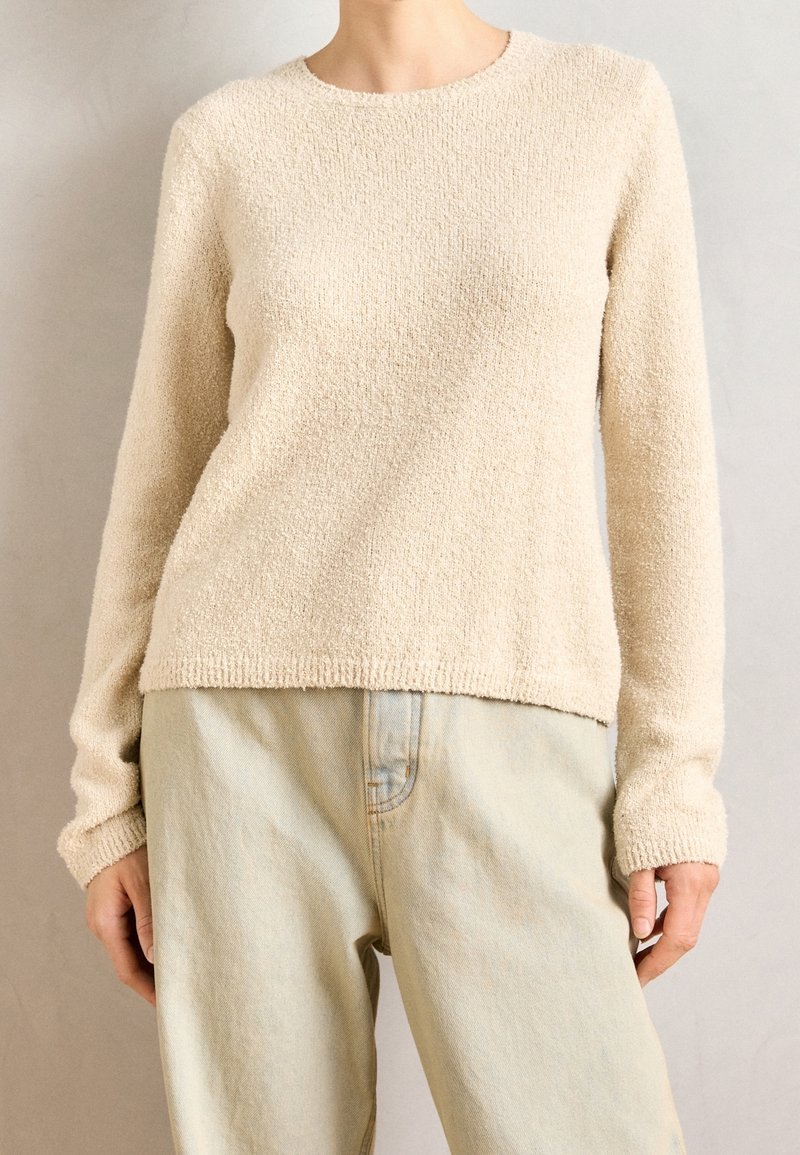 Personne portant un pull beige texturé à manches longues et un pantalon beige clair taille haute, debout devant un fond uni.