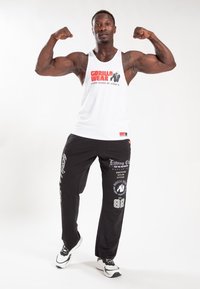 Balts bezmugurs tērps ar "GORILLA WEAR" tekstu; melni treniņbikses ar logotipiem un tekstu; sporta apavi. Modelis izstiepj rokas pret vienkāršu fonu.