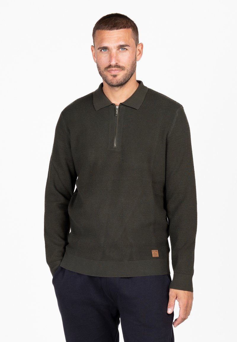 Dunkelgrüner Strickpullover mit einem Reißverschlusskragen, der eine horizontale Rippstruktur aufweist und ein kleines, geprägtes Lederpatch am Saum trägt.