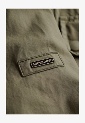 Olivengrønt stof med et syet mærke med "SUPERDRY" logoet i sort og guld, med en glat tekstur og afslappet design.