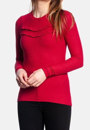 Vrouw draagt een nauwsluitend rood topje met lange mouwen en twee horizontale ruches over de borst, gecombineerd met een zwarte broek, handen in elkaar geklemd.