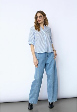 Femme portant un chemisier bleu clair à fines rayures, un jean ample délavé, des bottes noires à bout pointu et des lunettes, debout devant un fond blanc.