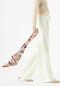 Foulard en soie avec un motif floral et abstrait en crème, bordeau et rose, tenu par une personne portant un pull beige clair et un pantalon blanc à jambes larges.