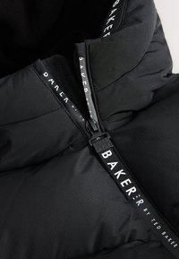 Gros plan sur la fermeture éclair d'une doudoune noire avec le texte « Baker by Ted Baker » sur la tirette et la bande en tissu.