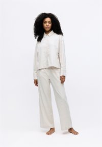 Lichtbeige, gemêleerd twee-delig outfit bestaande uit een korte, geknoopte blouse met kraag en wijde broekspijpen. Model staat op blote voeten.
