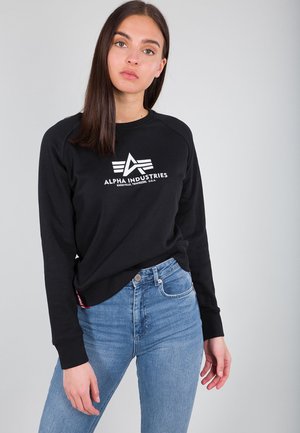 Black sweatshirt with white logo, crew neckline, and ribbed cuffs. Paired with light blue jeans, showcasing a casual fit and comfortable design.