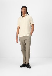 Lichtbeige korteblauwe shirt met een gestructureerde stof, met knopen; gecombineerd met lichtgrijze gestructureerde broek en zwarte schoenen.