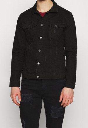 Mężczyzna w czarnej postarzanej kurtce dżinsowej na czerwonej koszulce i dopasowanych czarnych postarzanych dżinsach, stojący na białym tle.