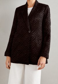 Blazer nero con un motivo a strisce zebra testurizzate, chiusura con un solo bottone, rever a punta e due tasche frontali. Indossato su una maglietta bianca.