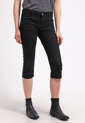 Kobieta ubrana w czarne krótkie spodnie typu slim fit, szarą wsuniętą koszulę i czarne botki, stojąca na białym tle.