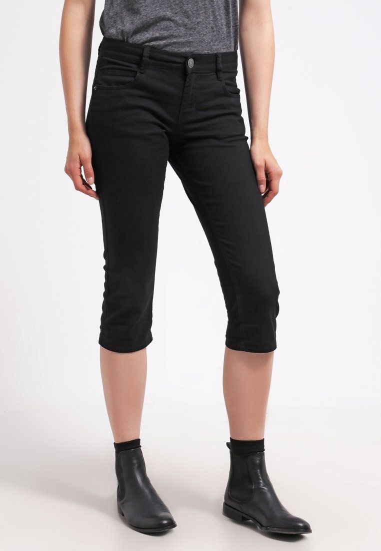 Femme portant un pantalon noir slim court, une chemise grise rentrée et des bottines noires, debout devant un fond blanc.