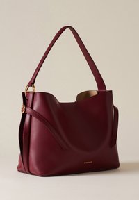 Borsa a mano in pelle bordeaux con corpo strutturato e morbido, forma affusolata e tracolla regolabile. Presenta cuciture sottili e un accento con logo dorato.