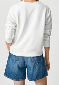 Felpa bianca in cotone con maniche lunghe, scollatura tonda e polsini a coste, abbinata a pantaloncini di jeans blu con tasche posteriori e orli sfrangiati.