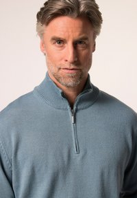 Světle modrý pletený svetr s vysokým límcem a předním zipem. Materiál je měkký a strukturovaný, s žebrovanými detaily na límci.