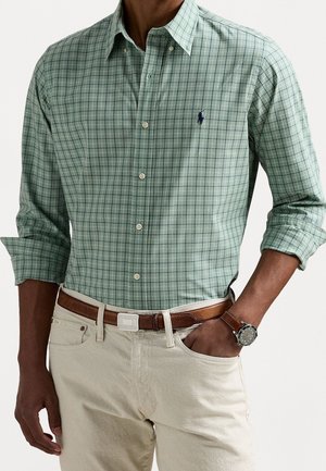 Mand iført lys grønternet skjorte med knapper og op-rullede ærmer, beige bukser, brun bælte og armbåndsur med brun læderrem.