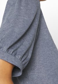 Camiseta gris texturizada con mangas cortas abullonadas, que presenta sutiles acentos de brillo. La tela es suave y ligera.