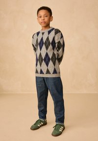 Szary sweter w argyle z granatowymi i czarnymi diamentami, niebieskie dżinsy oraz zielone sneakersy z białymi paskami. Proste, teksturowane tło.